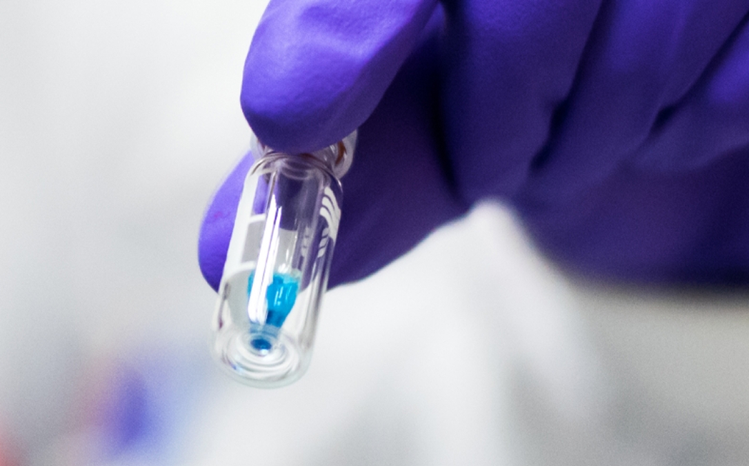 Eine behandschuhte Laborhand haelt ein Glasflaeschchen mit einem blauen Wirkstoff